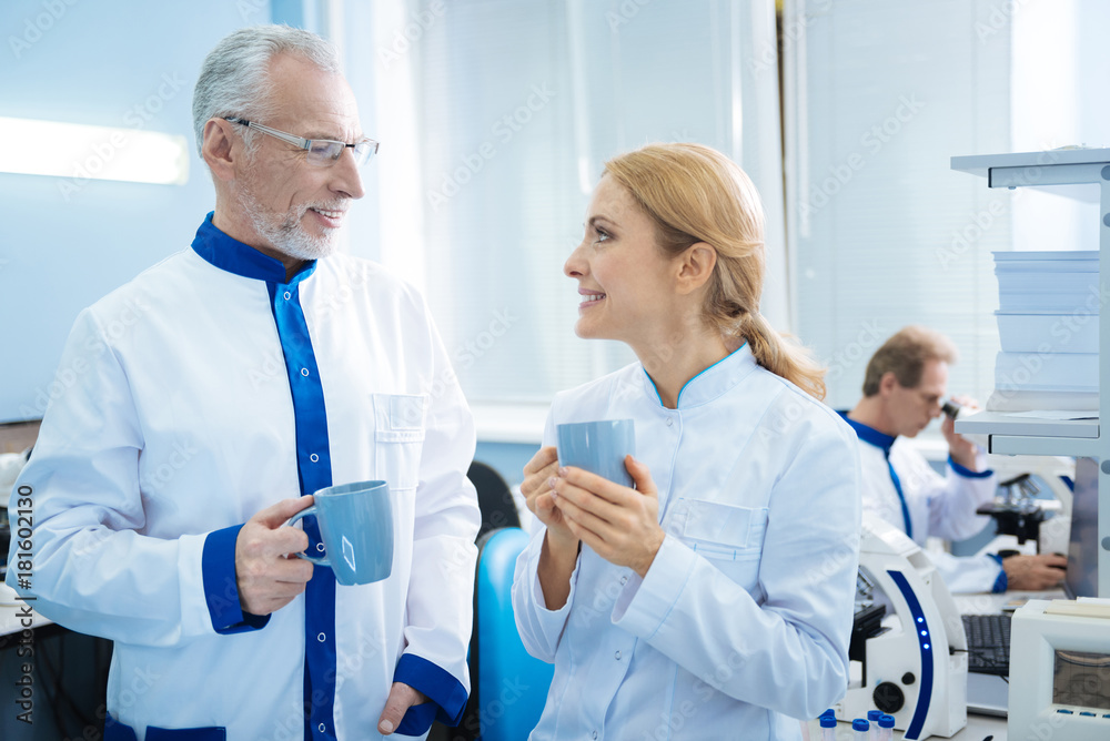 Coffee break. Exuberant smiling grey-haired bearded scientist and happy female blond researcher holding cups and looking at each other in the lab and one more researcher working in the background Coffee break. Exuberant smiling grey-haired bearded scientist and happy female blond researcher holding cups and looking at each other in the lab and one more researcher working in the background