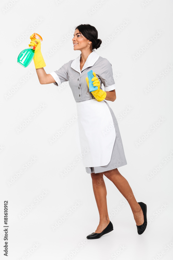 Full length photo of young brunette mais in uniform and yellow protective gloves spraying the cleaner on window Full length photo of young brunette mais in uniform and yellow protective gloves spraying the cleaner on window