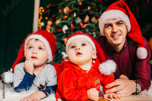 Two pretty little boys lie with their father on fluffy carpet before a Christmas tree