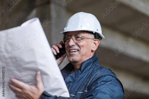 Senior construction manager controlling building site with plan and talking at phone.