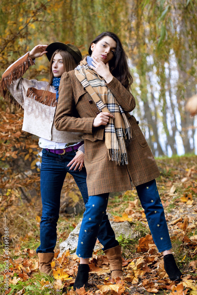 Fashion portrait of two beautiful women standing on grass. Autumn landscape. Trendy indian coat and hat with scarf. Fashion autumn concept.Closeup.