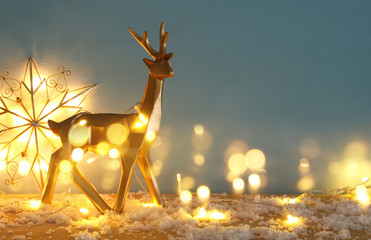 Gold shiny reindeer on snowy wooden table with christmas garland lights.