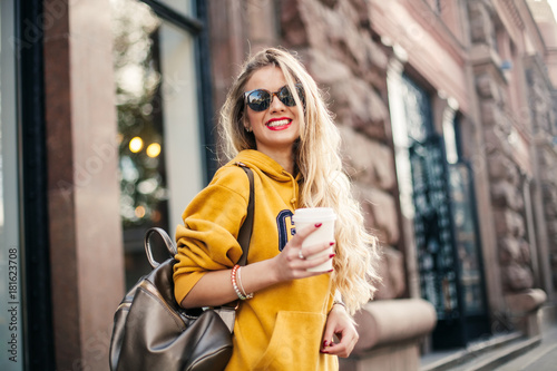 young beautiful blonde drinking coffee walking around the city.mustard sweetshot.,urban backpack , bright red lips Posing against the window of the boutique