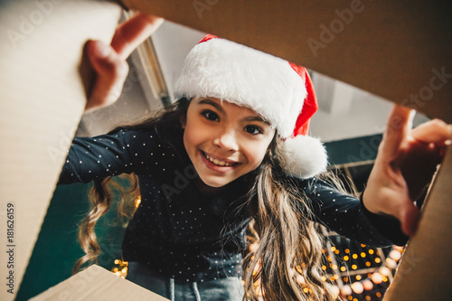 Surprised cute child girl opening a Christmas present. Little kid having fun near decorated tree indoors.  Happy  holidays and New Year.