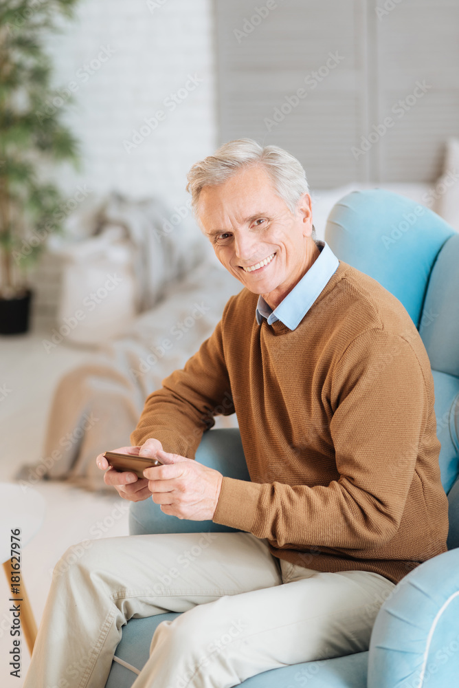 Enjoying my retirement. Radiant elderly gentleman looking into the camera with a cheerful smile on his face while sitting in a comfy chair and using his phone at home.