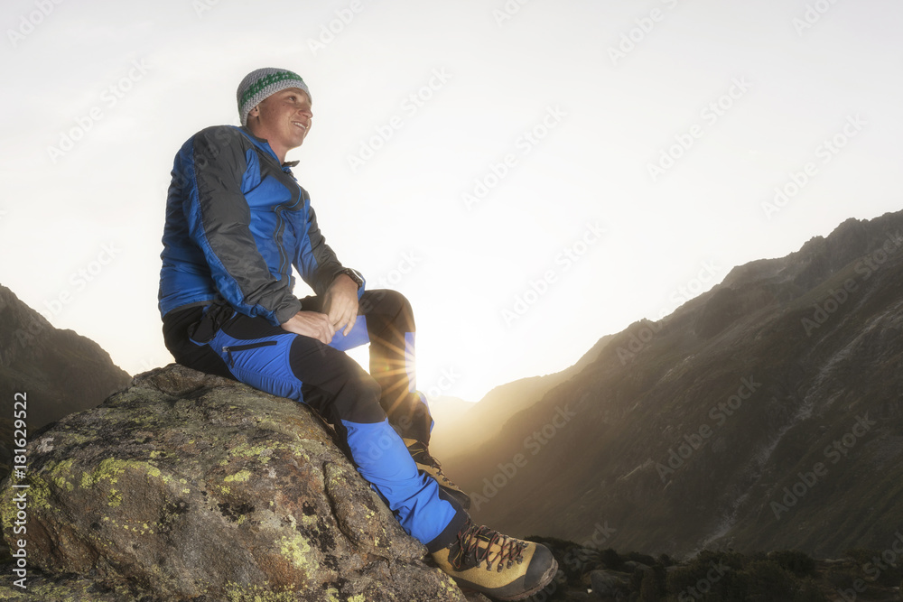 Good-looking young hiker taking a break on a stone and enjoys the sunrise in the swiss mountains Good-looking young hiker taking a break on a stone and enjoys the sunrise in the swiss mountains