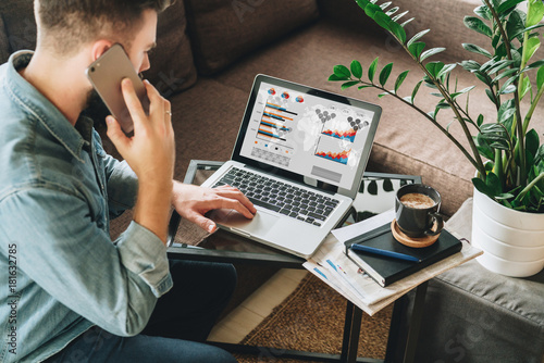 Photos Young man, dressed in shirt, sits at home on couch at coffee table,uses laptop with graphs, charts, diagrams on screen, talks on his cell phone,calling