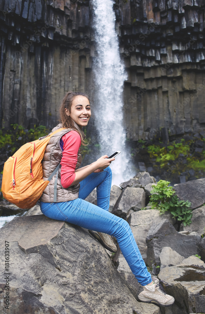 Woman tourist posing with smartphone on background of Svartifoss waterfall surrounded by basalt columns in the south of Iceland. Skaftafell national park in Iceland Woman tourist posing with smartphone on background of Svartifoss waterfall surrounded by basalt columns in the south of Iceland. Skaftafell national park in Iceland