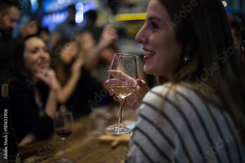 Young woman enjoys glass of dry white wine, chats and talks with friends at party or meeting in downtown alcohol bar, hipster beautiful going out style, open for communication and flirt