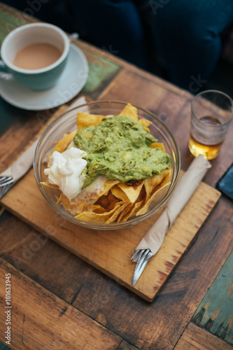 Top view of big bowl of crispy corn nachos or tortillas with sour cream and avocado spread or guacamole served on authentic artisan wooden cutting board in cool hipster snack place