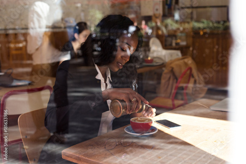 Beautiful young african american woman with long curly hair drinks coffee at sowntown cafe spot, adds brown sugar to hot caffeinated drink, casual lifestyle photo through glass