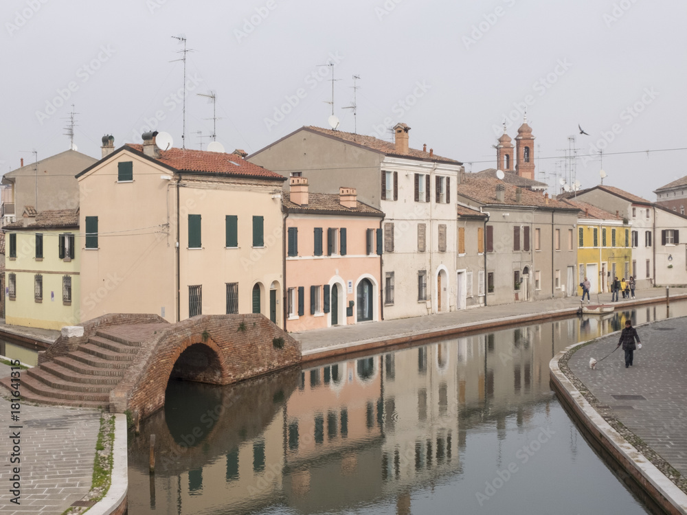 Comacchio, FE, Italy - November 4, 2017: view of one of the canals crossing Comacchio. The colored facades of the houses are reflected on the water. On one side of the many bridges on the canal. Comacchio, FE, Italy - November 4, 2017: view of one of the canals crossing Comacchio. The colored facades of the houses are reflected on the water. On one side of the many bridges on the canal.