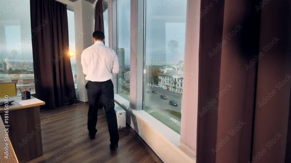 A businessman comes to stand near an office window to work. A businessman comes to stand near an office window to work.