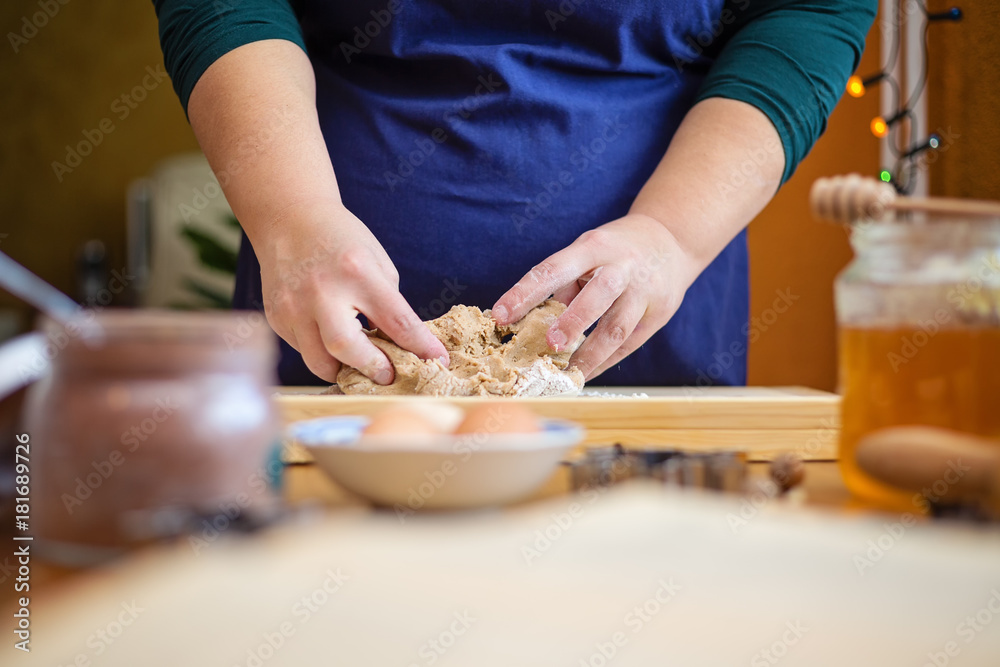 Close up of young woman hands kneading a dough on a wooden board. She is standing in the kitchen, behind a table, next to a window decorated with christmas lights