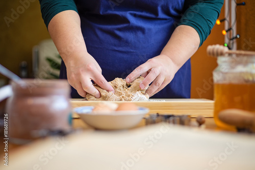 Close up of young woman hands kneading a dough on a wooden board. She is standing in the kitchen, behind a table, next to a window decorated with christmas lights