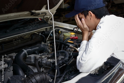 Frustrated stressed young Asian mechanic man in white uniform covering face with hands against car in open hood at the repair garage.