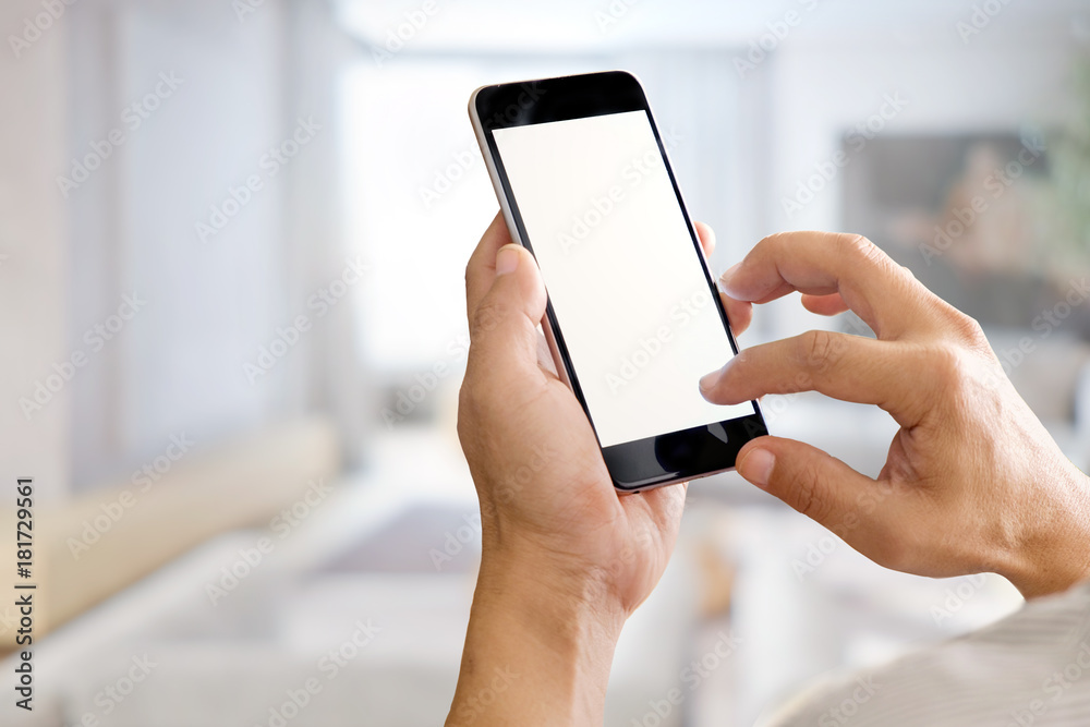 Mockup image of hands holding black mobile phone with blank white screen in living room.