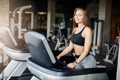 Portrait of crazy fit woman on a threadmill smiling looking at camera. Athletic coach warming up early in the morning in empty gym.