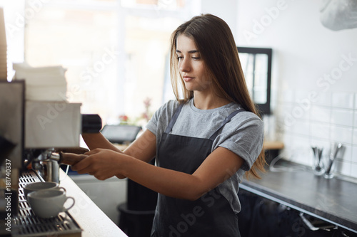Young and sleepy female barista concentrated on using a coffee machine to brew some fresh ground beans to wake up.