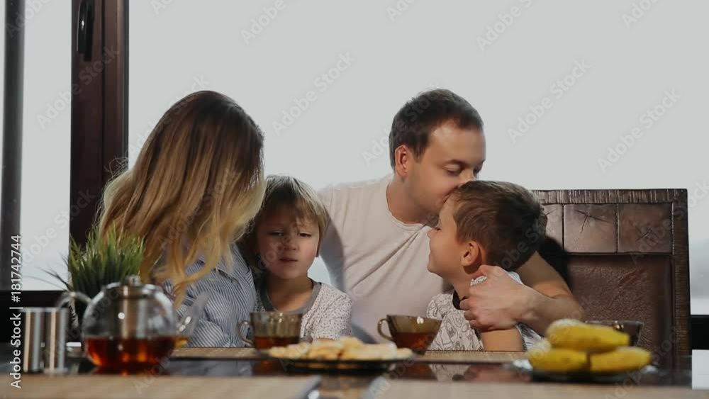 Family of four having Breakfast in his kitchen with large Windows. People are smiling, mother kissing and hugging children. Mother father and two children. Family of four having Breakfast in his kitchen with large Windows. People are smiling, mother kissing and hugging children. Mother father and two children.