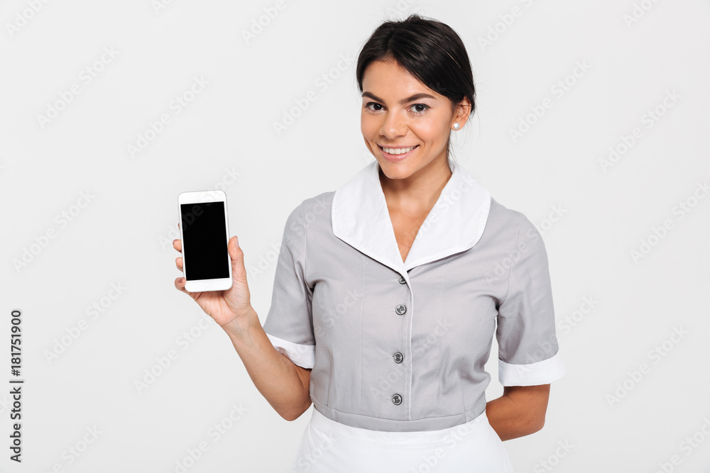 Cheerful brunette girl in gray uniform showing blank mobile screen while standing and looking at camera Cheerful brunette girl in gray uniform showing blank mobile screen while standing and looking at camera