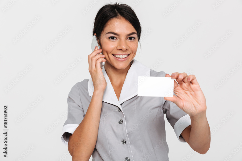 Portrait of young smiling brunette woman in uniform talking on mobile phone while showing empty sign card Portrait of young smiling brunette woman in uniform talking on mobile phone while showing empty sign card