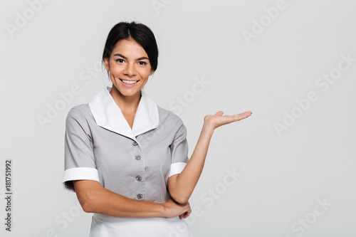 Close-up portrait of young happy housekeeper in uniform showing empty palm while looking at camera
