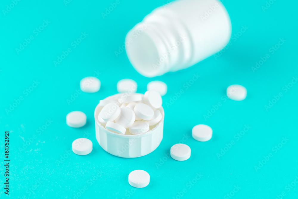 Close up white pill bottle with spilled out pills and capsules in cap on turquoise background with copy space. Focus on foreground, soft bokeh. Pharmacy drugstore concept Close up white pill bottle with spilled out pills and capsules in cap on turquoise background with copy space. Focus on foreground, soft bokeh. Pharmacy drugstore concept