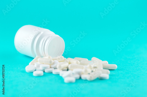 Close up white pill bottle with spilled out pills and capsules on turquoise background with copy space. Focus on foreground, soft bokeh. Pharmacy drugstore concept