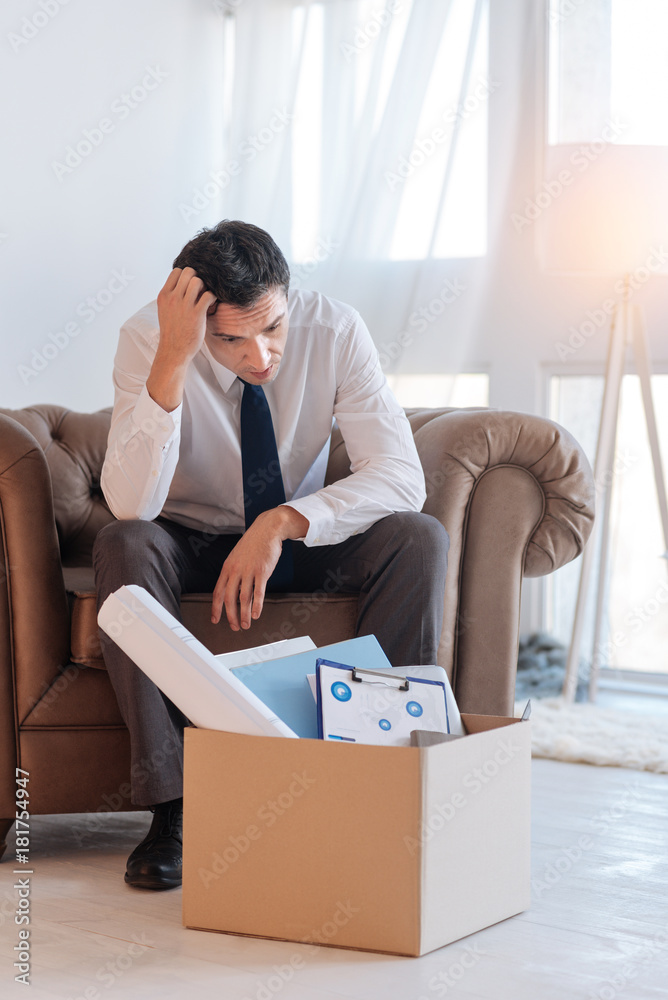 Strange. Curious thoughtful young man sitting in a comfortable brown armchair while scratching his head and looking attentively at the big box on the floor
