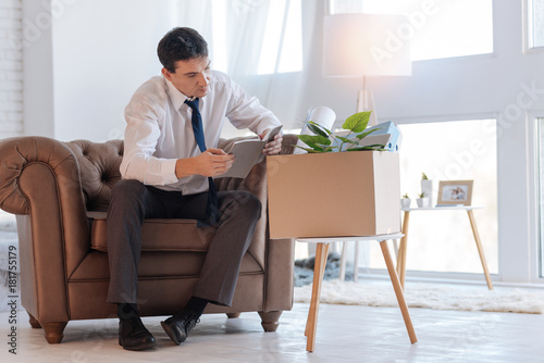 Reading. Attentive concentrated jobless man sitting in a brown armchair and reading old notes in a notebook while a box with personal things standing on a chair by his side