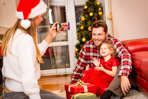 christmas, holidays, family and people concept - mother photographing happy father and little daughter with present by smartphone