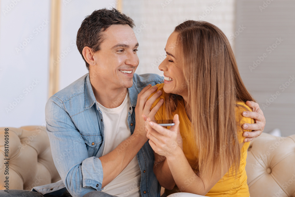 Great device. Excited cheerful young woman feeling happy while holding a new convenient smart phone while her beloved man sitting by her side and hugging her Great device. Excited cheerful young woman feeling happy while holding a new convenient smart phone while her beloved man sitting by her side and hugging her