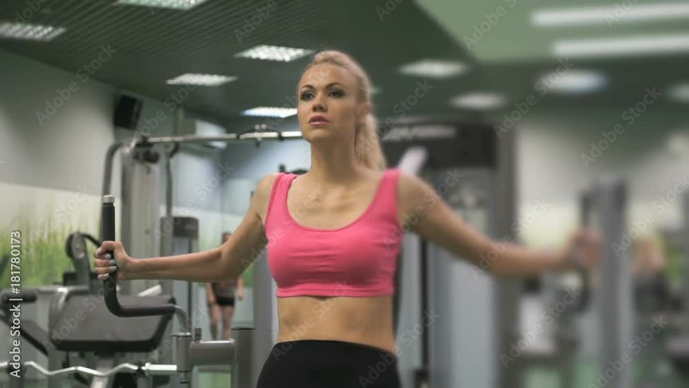 Sports workout in the gym indoors. Young beautiful blond girl in sport suit training her arms on the sports simulator