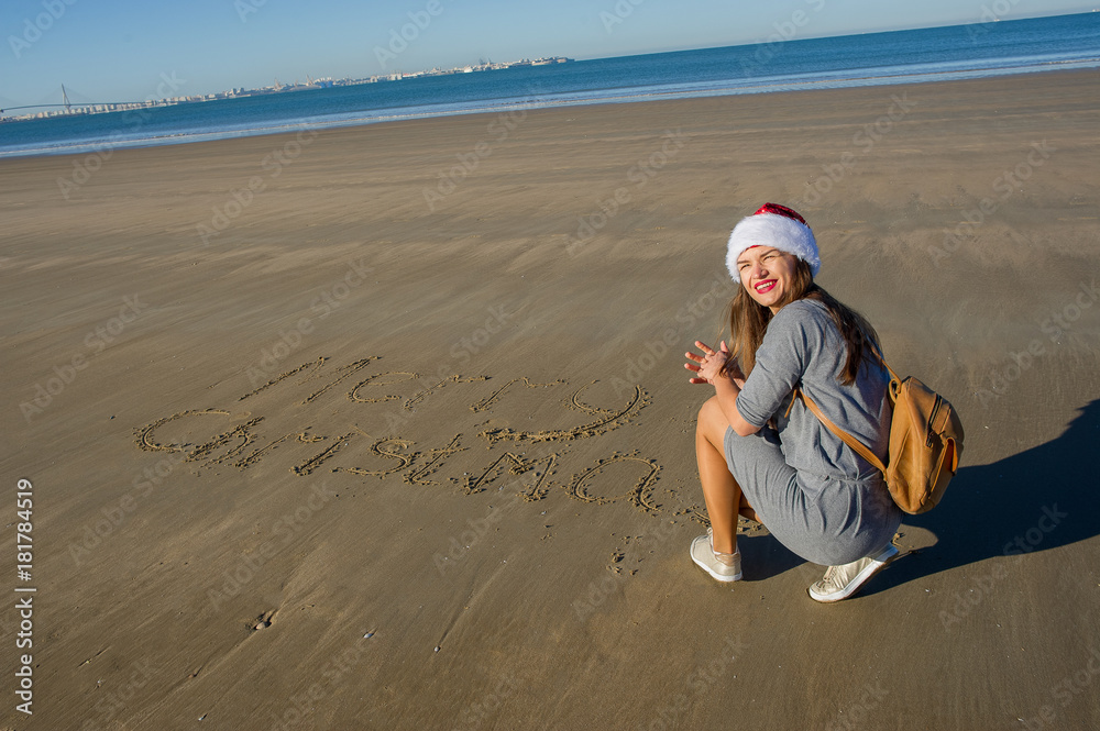 Merry Christmas! Charming girl in a Santa hat wrote greetings on the sand. Merry Christmas! Charming girl in a Santa hat wrote greetings on the sand.