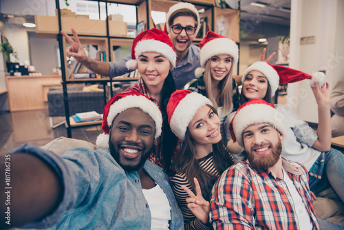 Self portrait of mixed race friends : african, american, asian, caucasian,  young smiling bearded men and beautiful women in red christmas hat showing peace symbol  to the camera