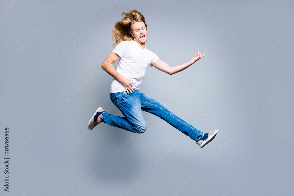 Music is my life! Excited and amazed young guy with blonde long hair in casual clothes is celebrating his victory, he is playing an imaginary guitar, isolated on grey background