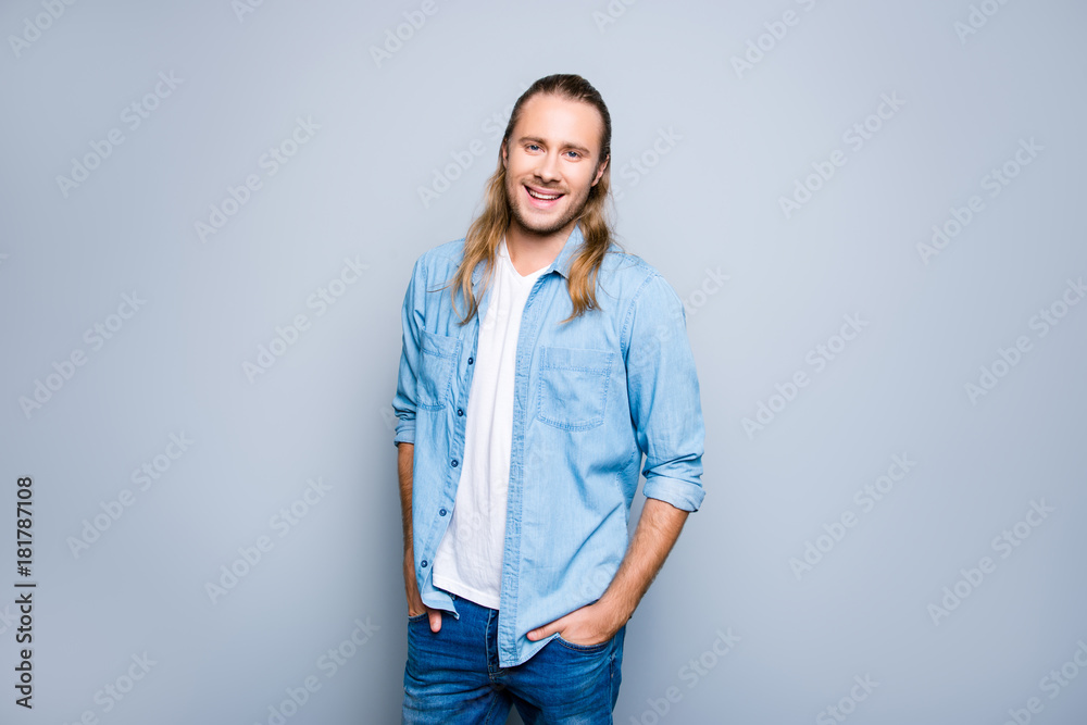Happy joyful young hipster with long blonde hair is smiling, holding his hands in pockets ans posing against grey background Happy joyful young hipster with long blonde hair is smiling, holding his hands in pockets ans posing against grey background