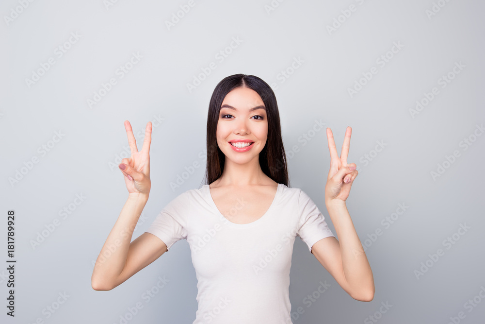 Portrait of glad, funny, smiling, toothy, beautiful girl showing with two hands v-sign, two peace symbols, standing over grey background Portrait of glad, funny, smiling, toothy, beautiful girl showing with two hands v-sign, two peace symbols, standing over grey background