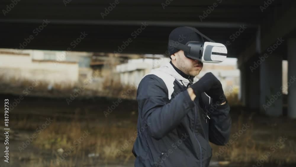 Tracking around shot of boxing man in VR 360 headset training punches in virtual reality fight at urban location outdoors in winter Tracking around shot of boxing man in VR 360 headset training punches in virtual reality fight at urban location outdoors in winter