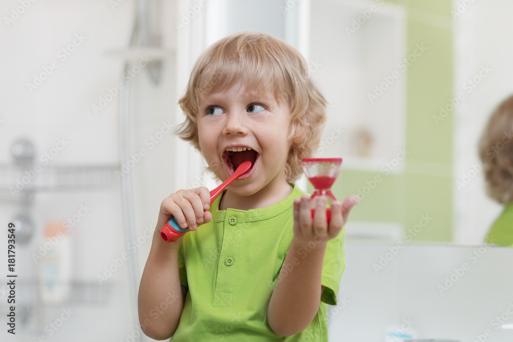Happy child brushing teeth near mirror in bathroom. He is monitoring lasting of cleaning action with hourglass. Happy child brushing teeth near mirror in bathroom. He is monitoring lasting of cleaning action with hourglass.