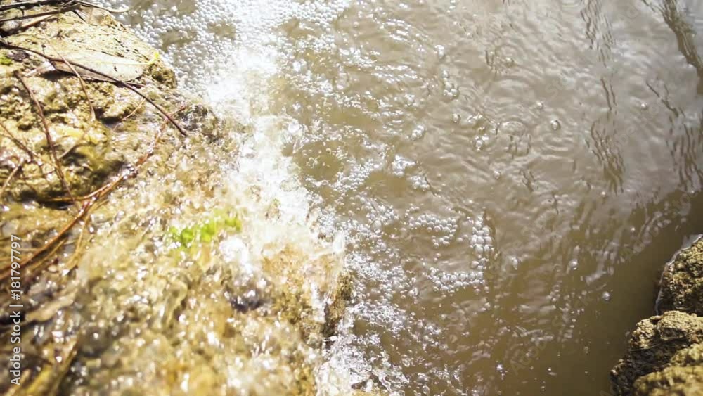Slow motion the spring stream of the stream drains down from the muddy shore with branches and leaves under the sun's rays top view Slow motion the spring stream of the stream drains down from the muddy shore with branches and leaves under the sun's rays top view