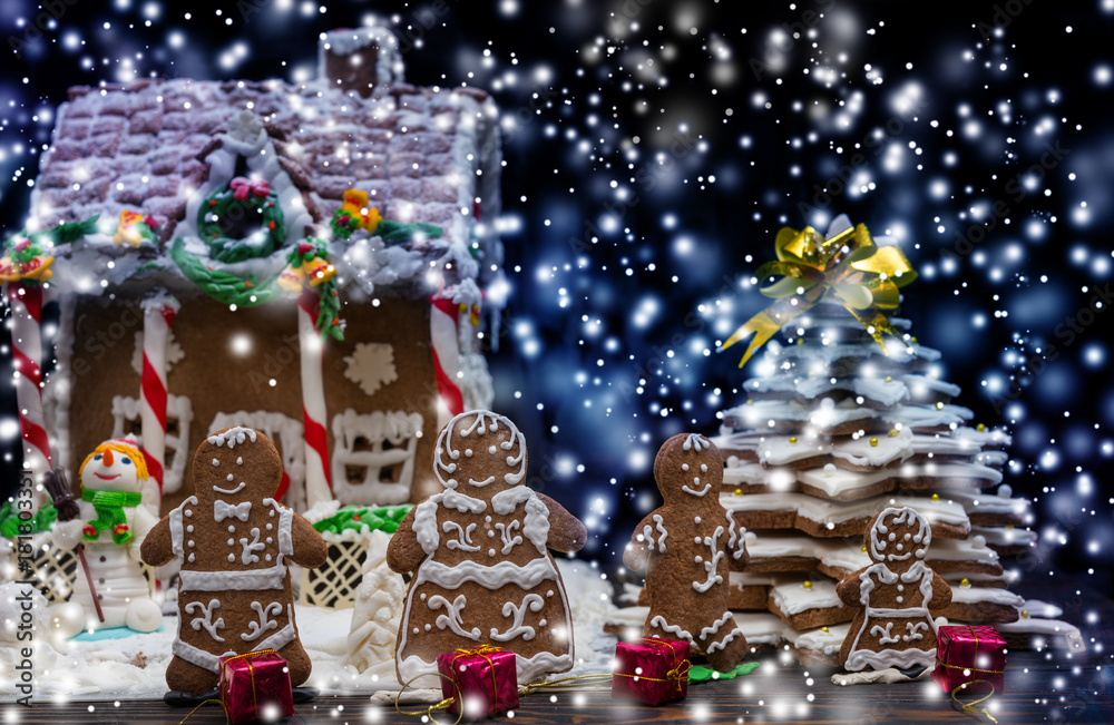 Cute gingerbread family with small red gifts near snow-covered homemade gingerbread house and Christmas tree during the snow
