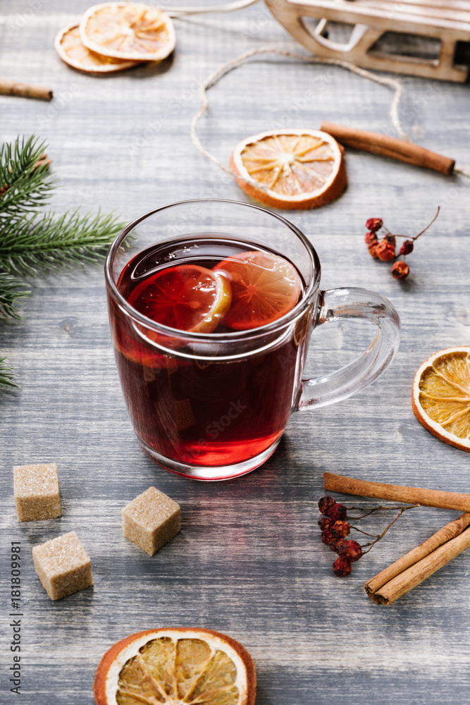 Hot berry tea with a lemon. Red herbal tea in glass cup on wooden grey table. Green fir-tree branch, dried orange, cinnamon Hot berry tea with a lemon. Red herbal tea in glass cup on wooden grey table. Green fir-tree branch, dried orange, cinnamon