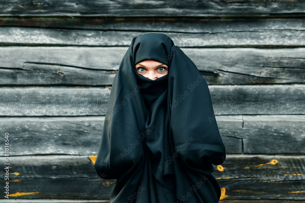 A beautiful, young, Muslim girl in a black veil with a closed face on a gray tree background holds her hands behind her head. Copy space.