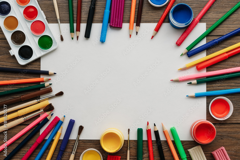 Naklejka premium A white sheet of A4 paper lies on a wooden brown table next to paints, brushes, colored pencils. View from above. The concept of creativity, creative, drawing.