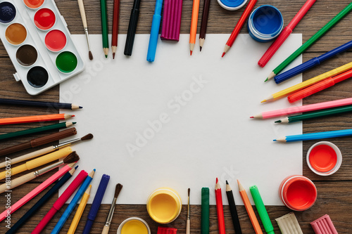 A white sheet of A4 paper lies on a wooden brown table next to paints, brushes, colored pencils. View from above. The concept of creativity, creative, drawing. photo