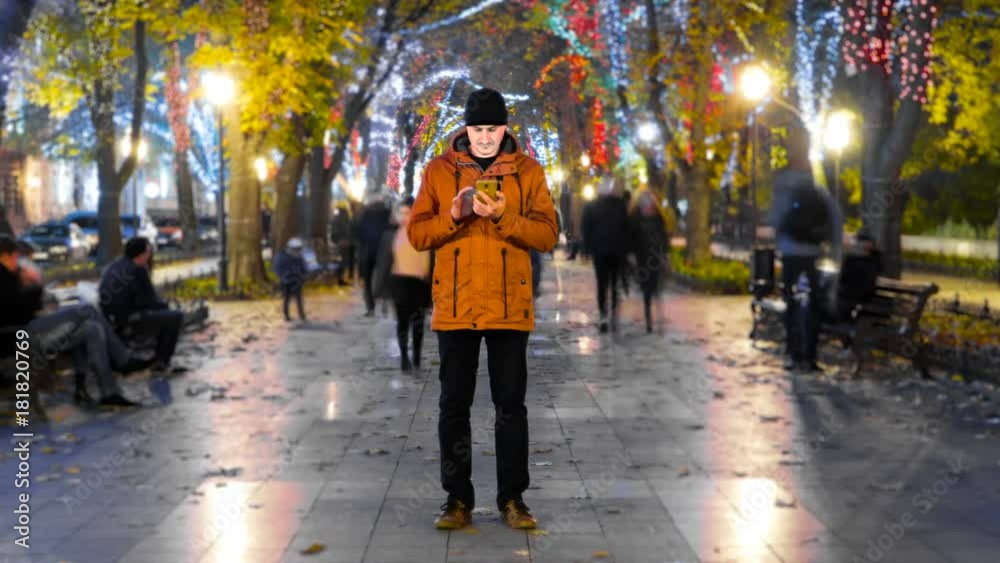 Timelapse of man standing still with smartphone on crowded walkway park boulevard with christmas garlands at night. People moving fast. Concept of flowing time and technologies Timelapse of man standing still with smartphone on crowded walkway park boulevard with christmas garlands at night. People moving fast. Concept of flowing time and technologies