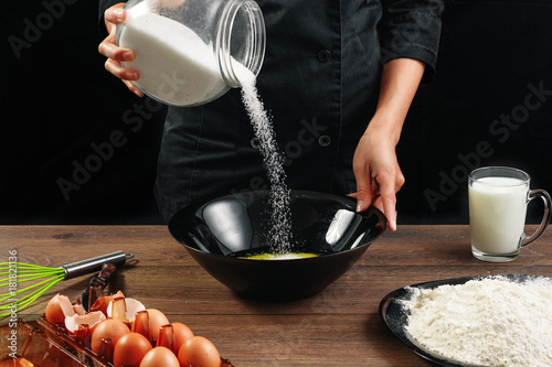 Male hands Chef chef pours sugar on a plate on a wooden brown table in black utensils. Chef kneads the dough. Black background. The concept of cooking food. Close-up. Copy space. photo