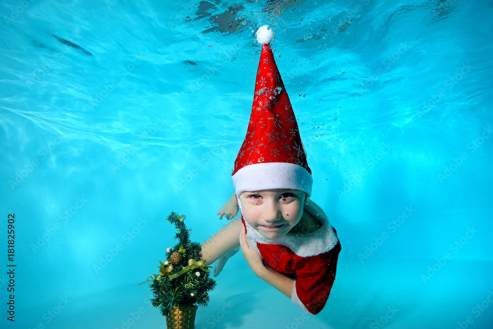 Happy child in a hat Santa Claus swims and poses underwater with a Christmas tree on a blue background. Portrait. Close-up. Horizontal orientation Happy child in a hat Santa Claus swims and poses underwater with a Christmas tree on a blue background. Portrait. Close-up. Horizontal orientation